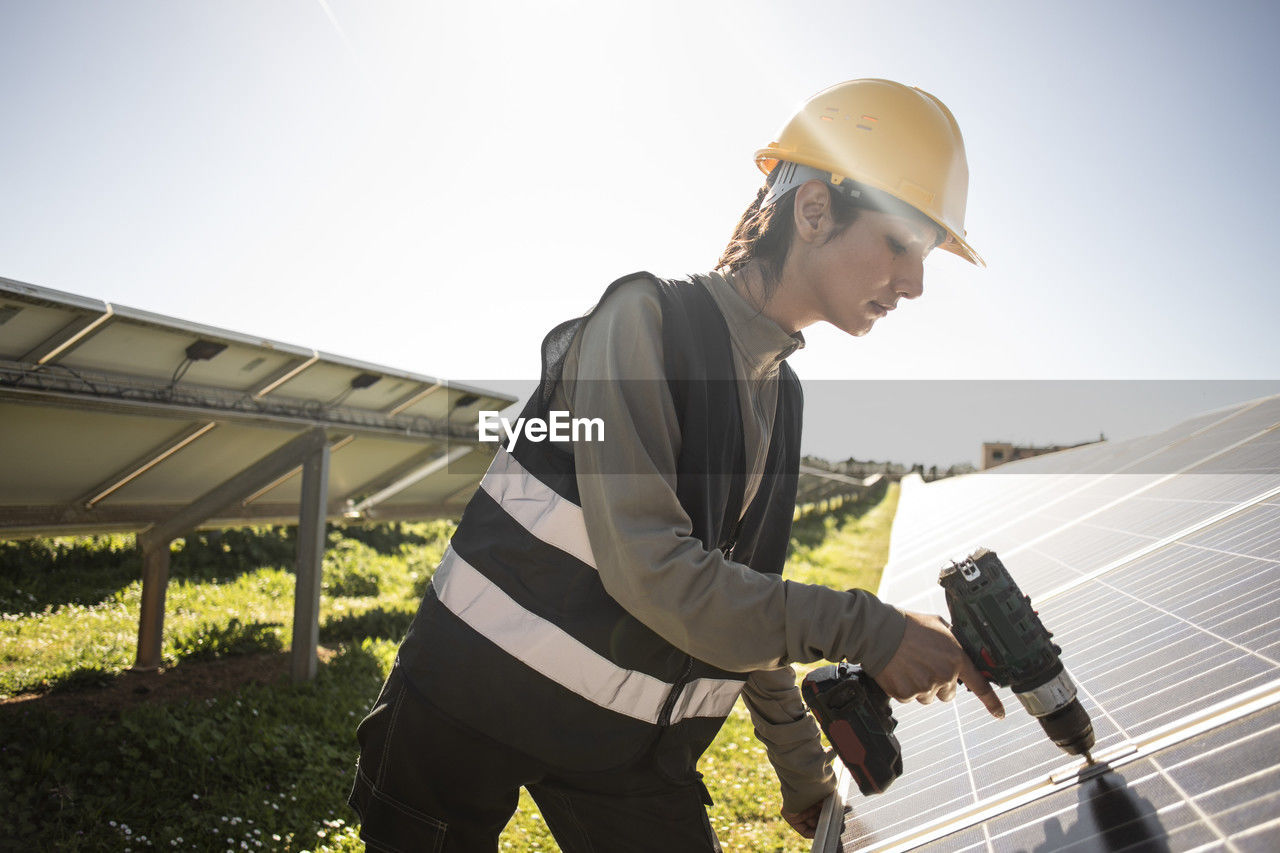 Female engineer using drill while working near solar panels at power station