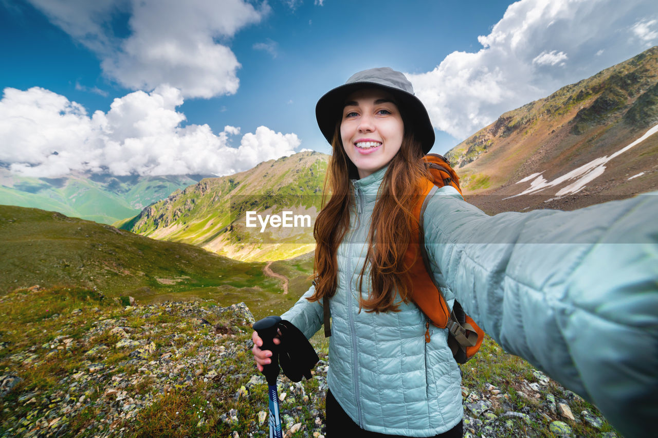 Smiling young woman takes a selfie on a mountain peak, with a backpack on a sunny day. tourist