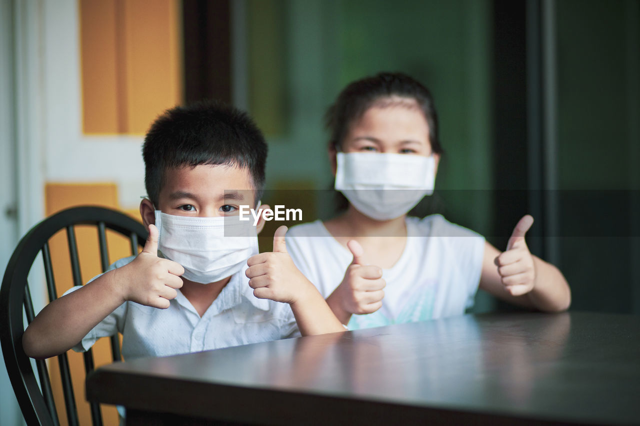 Portrait of sibling wearing mask gesturing while sitting by table at home