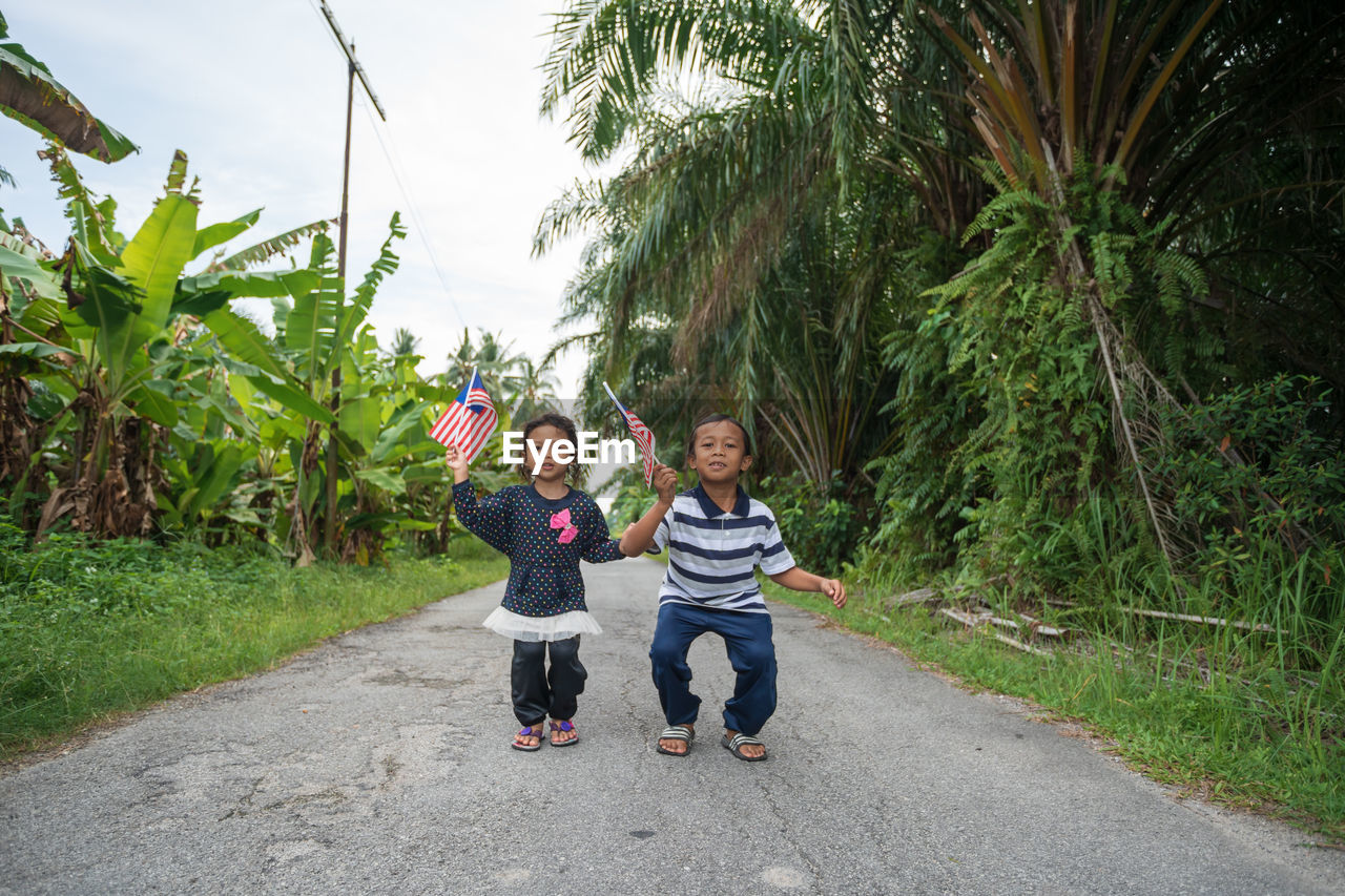 Siblings holding malaysian flag while standing on road amidst plants