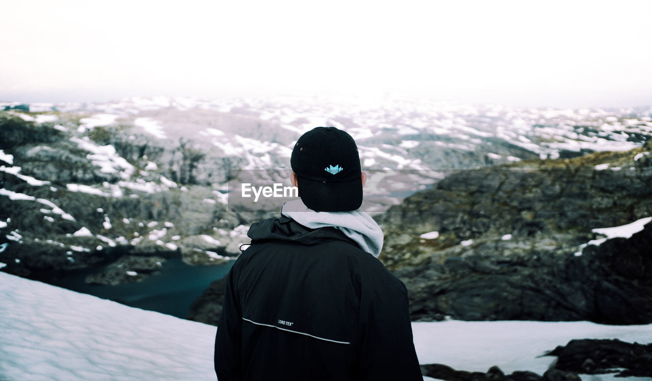 REAR VIEW OF MAN STANDING ON SNOW AGAINST MOUNTAIN