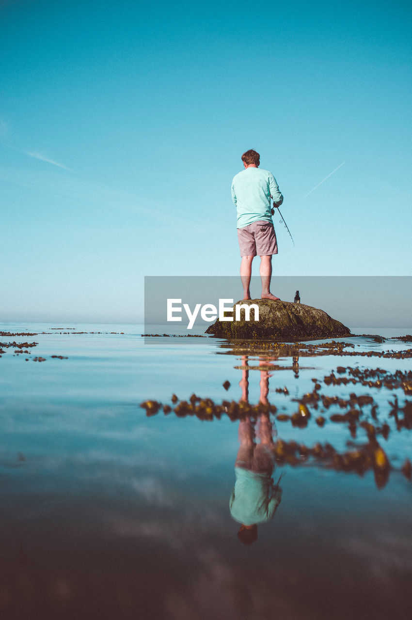 Rear view of man fishing at beach against blue sky