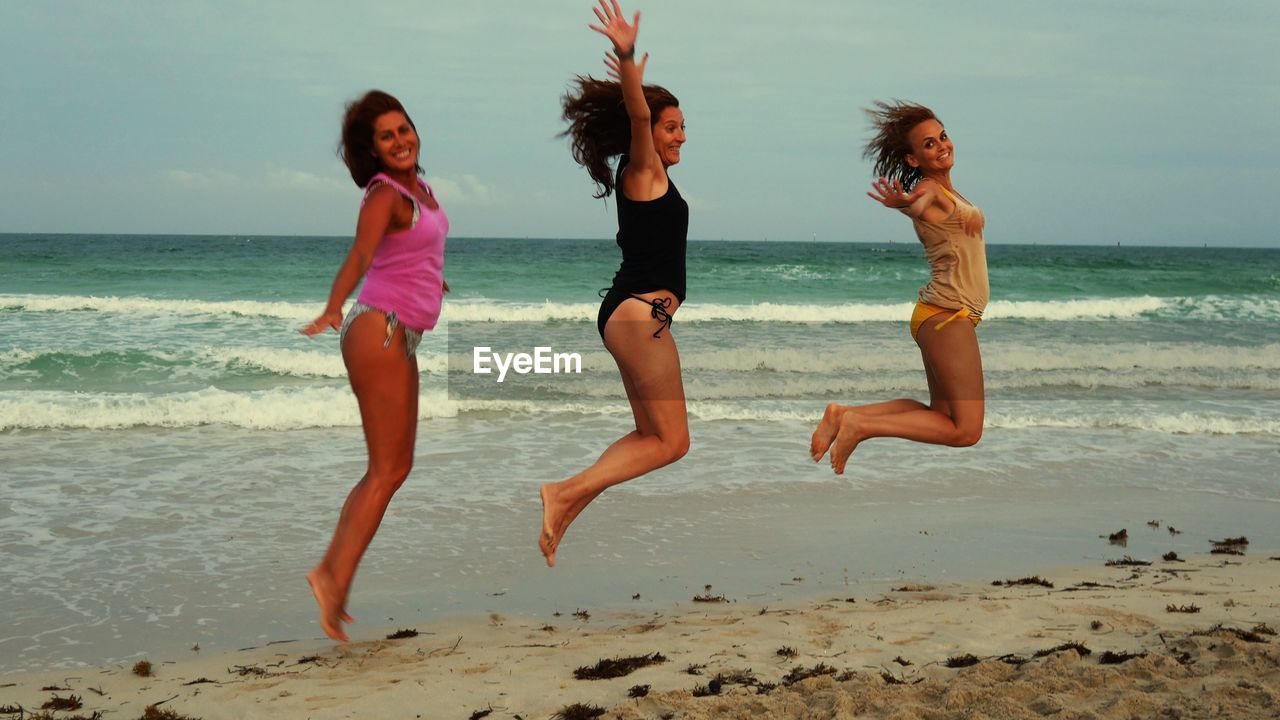 Happy friends jumping on sand against sea during sunset