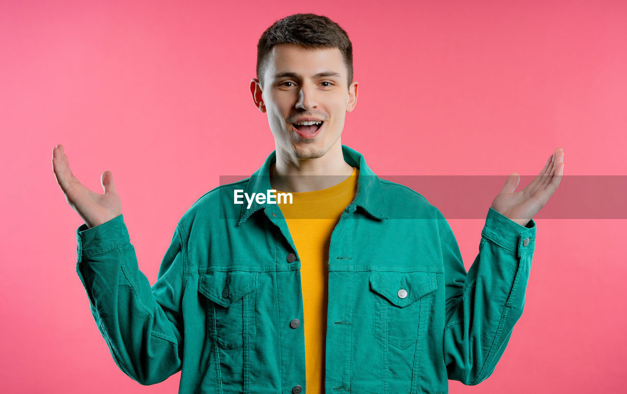 studio shot, one person, colored background, smiling, portrait, happiness, young adult, emotion, adult, waist up, looking at camera, cheerful, front view, indoors, men, positive emotion, gesturing, outerwear, standing, fun, clothing, green, hood, teeth, casual clothing, smile, person, jacket, pink background, pink, enjoyment, sleeve, singing, vitality, excitement, facial expression, hand sign, sign language, arm