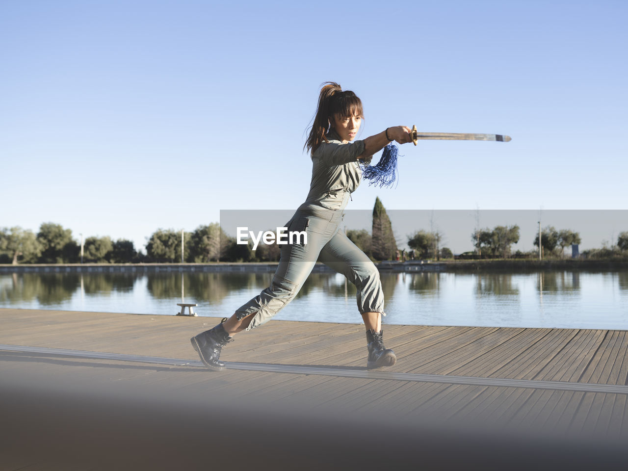 Female athlete practicing martial arts with sword by lake against clear sky