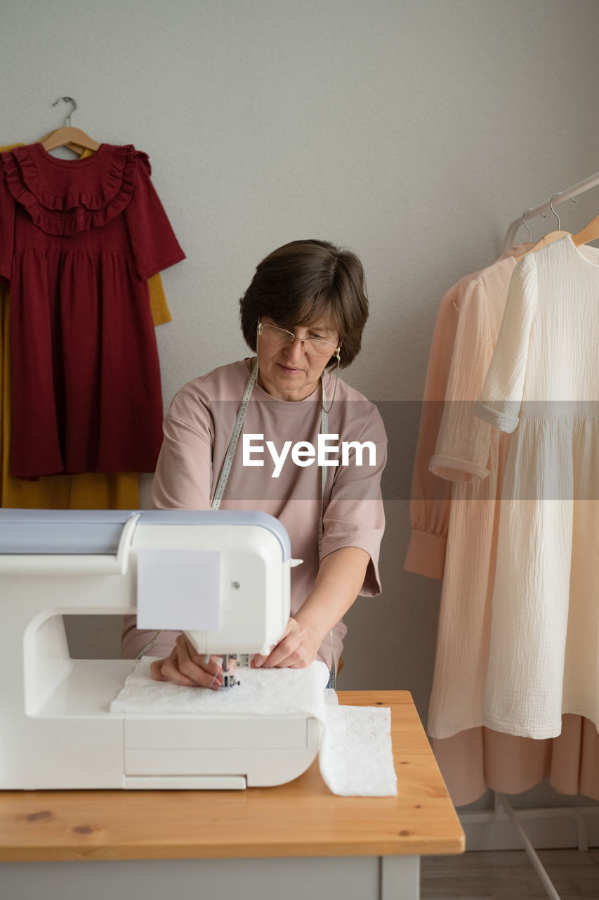 Concentrated female dressmaker in eyeglasses working on sewing machine at table with threads in light professional workshop with special equipment