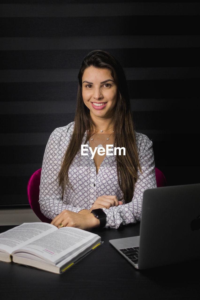 Portrait of smiling young woman with book and laptop on table sitting in office