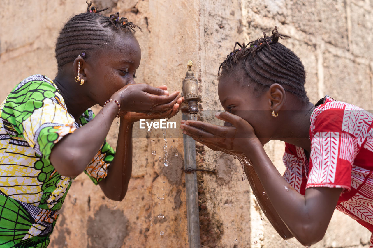 Girls drinking water from faucet against wall