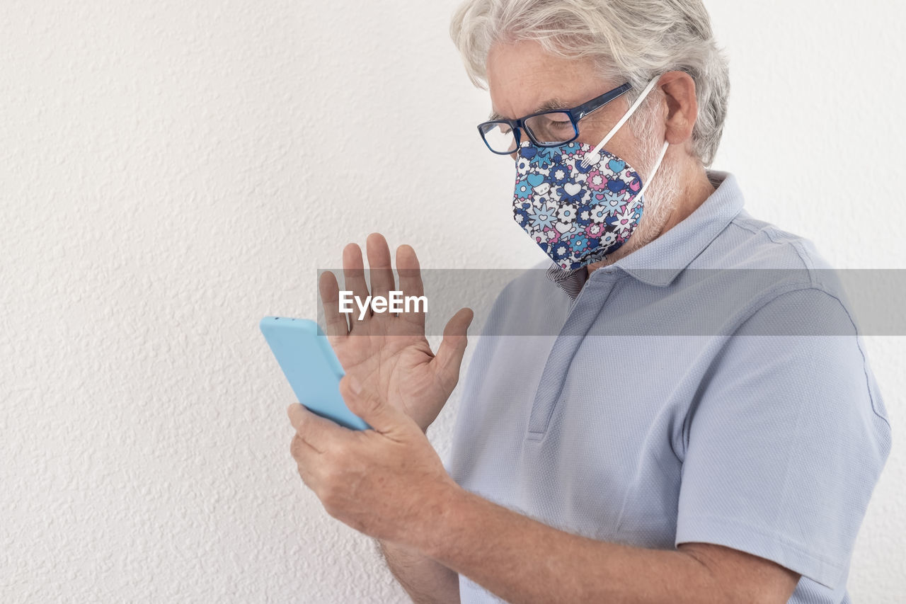 Senior man wearing mask using smart phone standing against wall