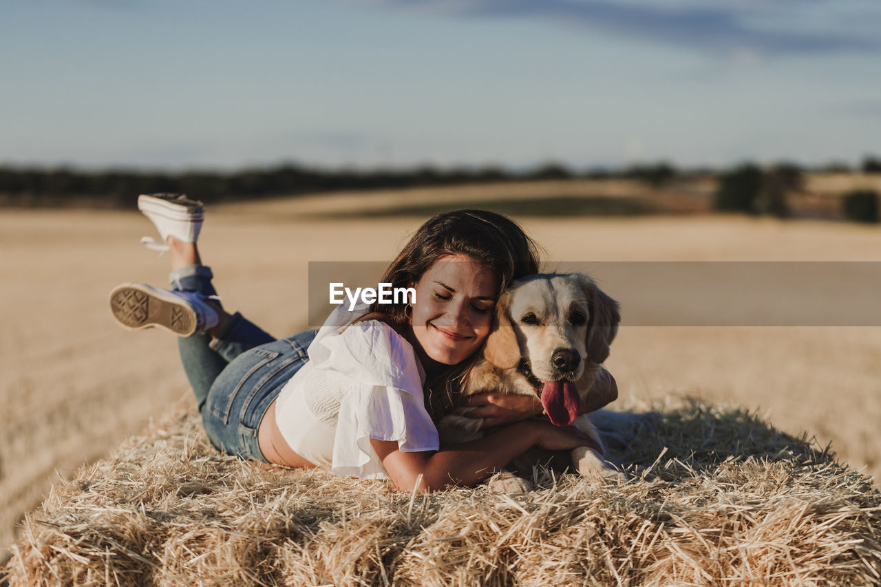 Happy mid adult woman with dog lying on hay at agricultural field