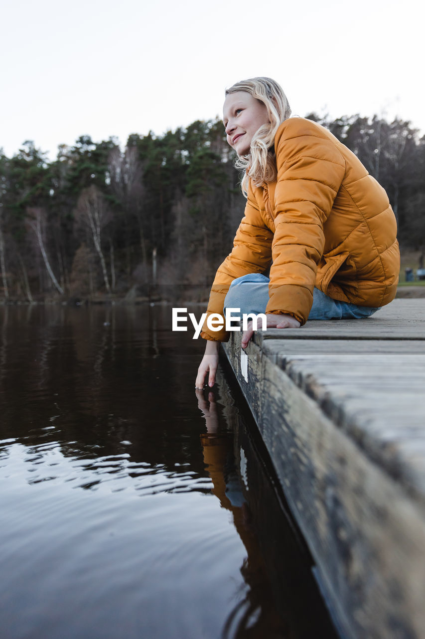 Low angle of cheerful teenage girl sitting on wooden quay near pond in autumn forest and looking away