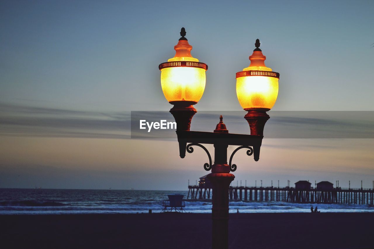 Illuminated street light by sea against sky during sunset