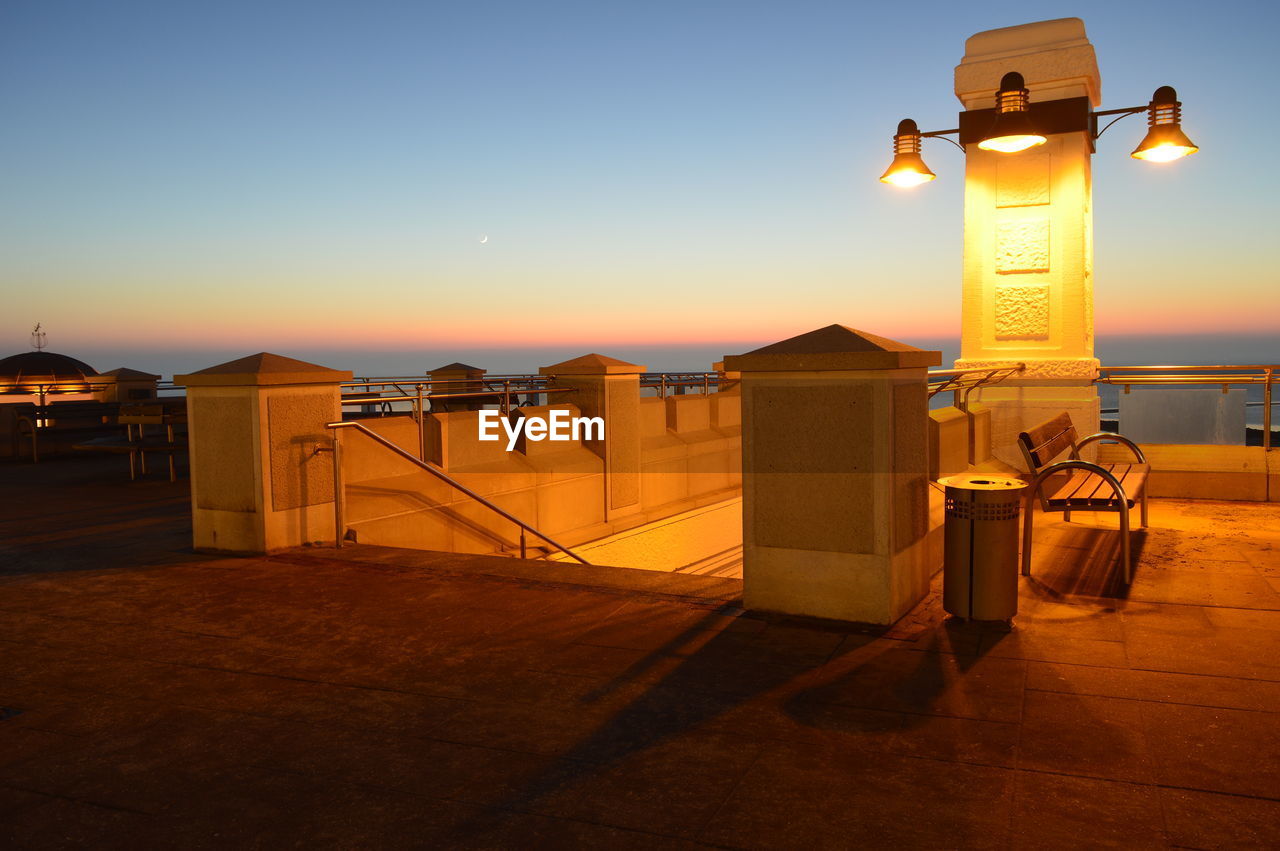 View of empty street by sea against clear sky at sunset