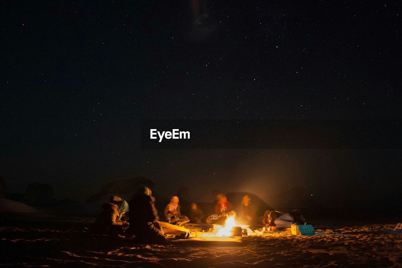 People sitting by bonfire against sky at night