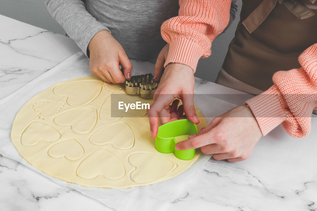 Two caucasian girls cut out shortbread dough with metal and plastic heart shapes.