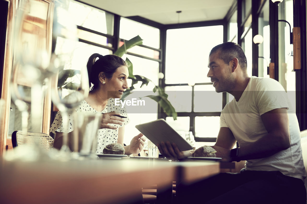 Low angle view of male and female with digital tablet talking while sitting at table in cafe