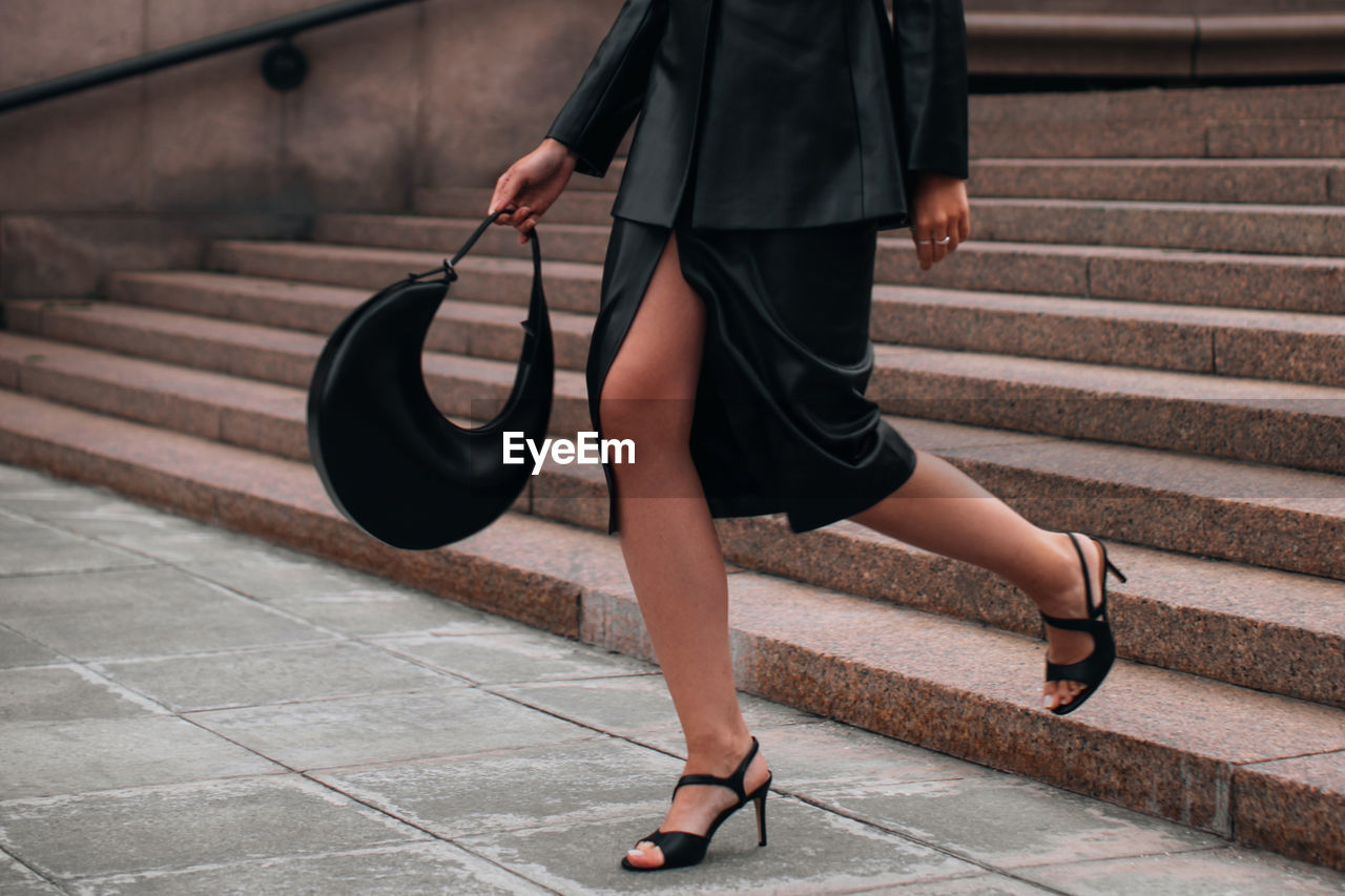 Fashionable details of a black leather skirt, jacket and handbag. woman walking in the city. 