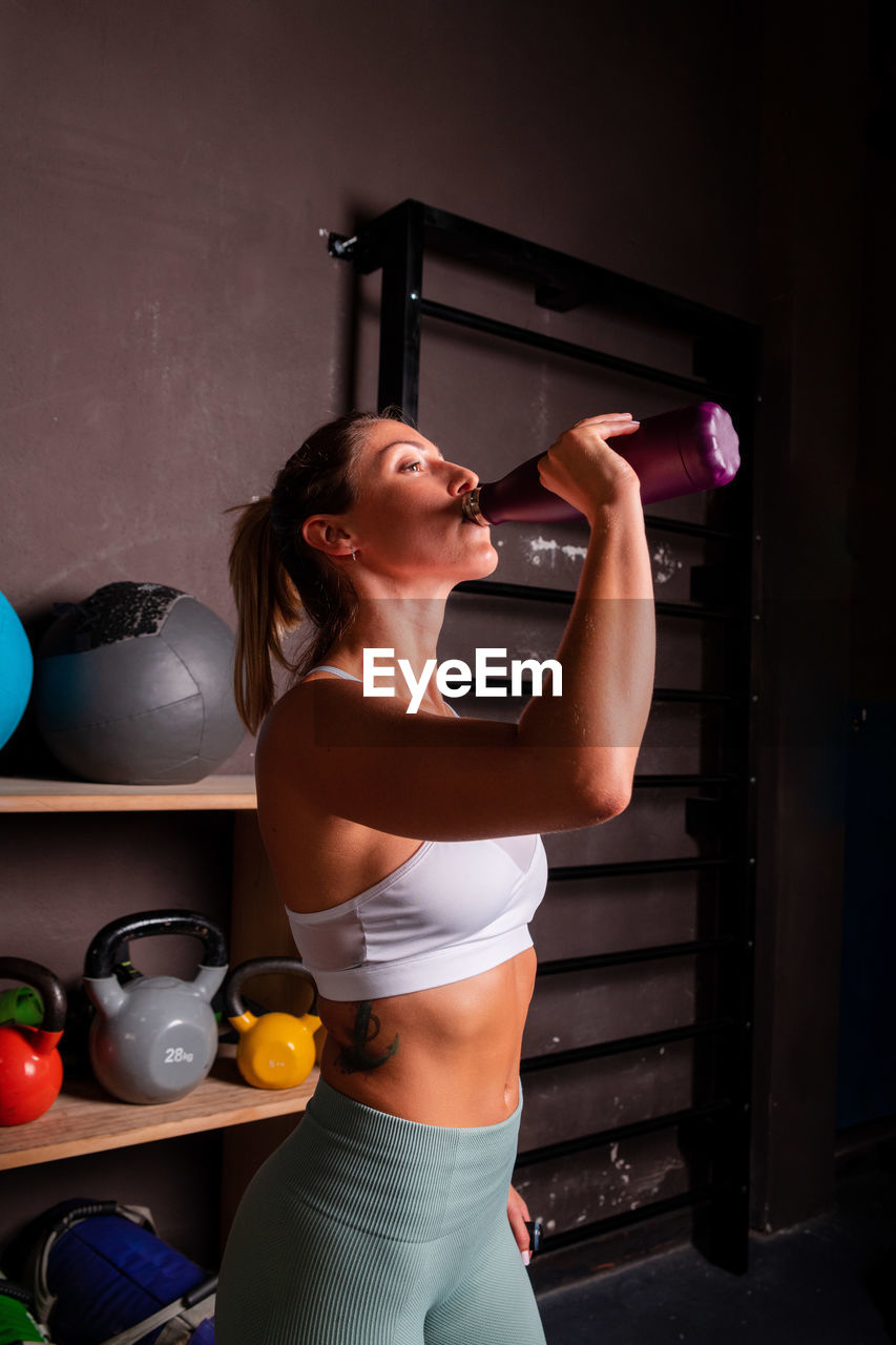 Side view of slim sportswoman in bra and leggings standing in modern gym and enjoying fresh water during training