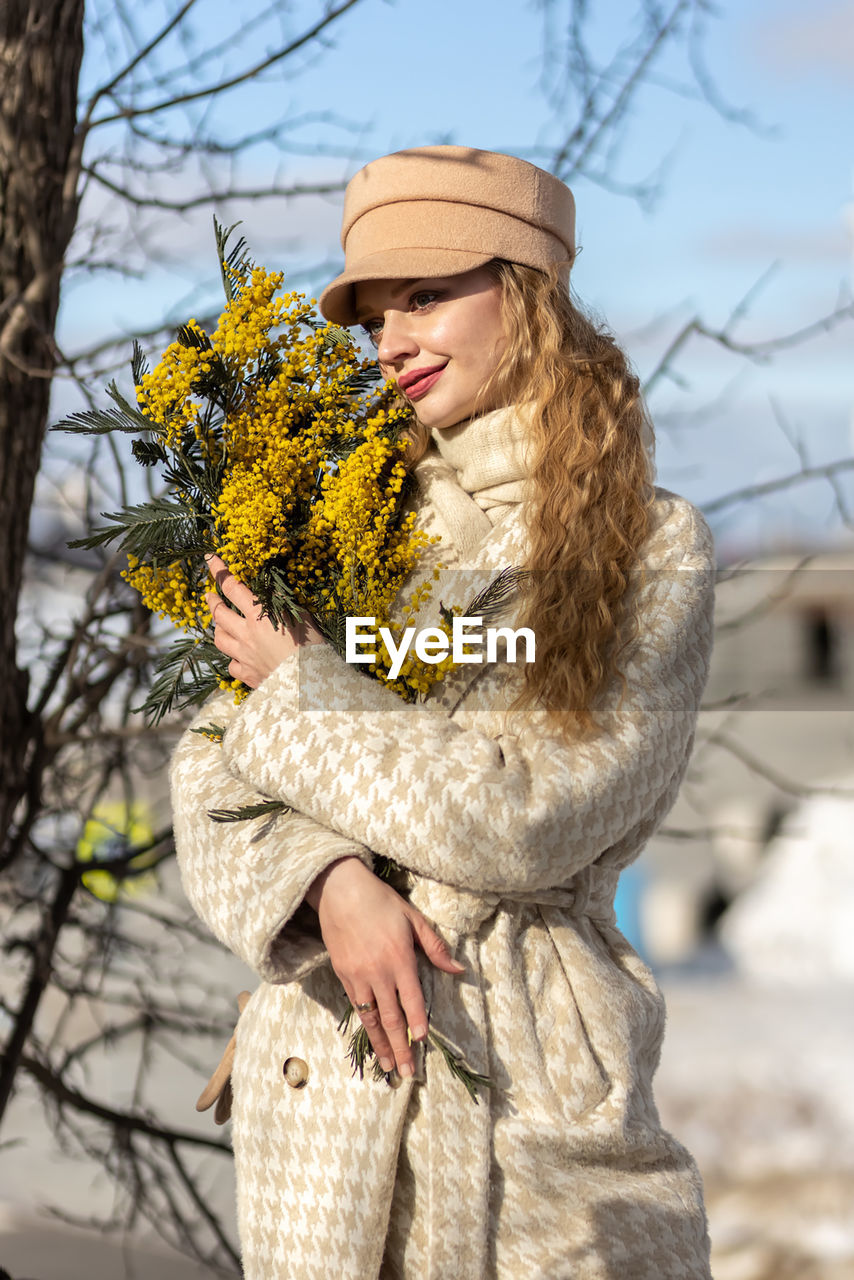 spring, clothing, adult, one person, women, flower, plant, winter, nature, young adult, warm clothing, hat, tree, flowering plant, long hair, cold temperature, fashion, hairstyle, emotion, portrait, beauty in nature, standing, smiling, holding, female, autumn, outdoors, happiness, looking, leisure activity, lifestyles, yellow, day, snow, blond hair, person, positive emotion, three quarter length, glove, focus on foreground, coat, front view