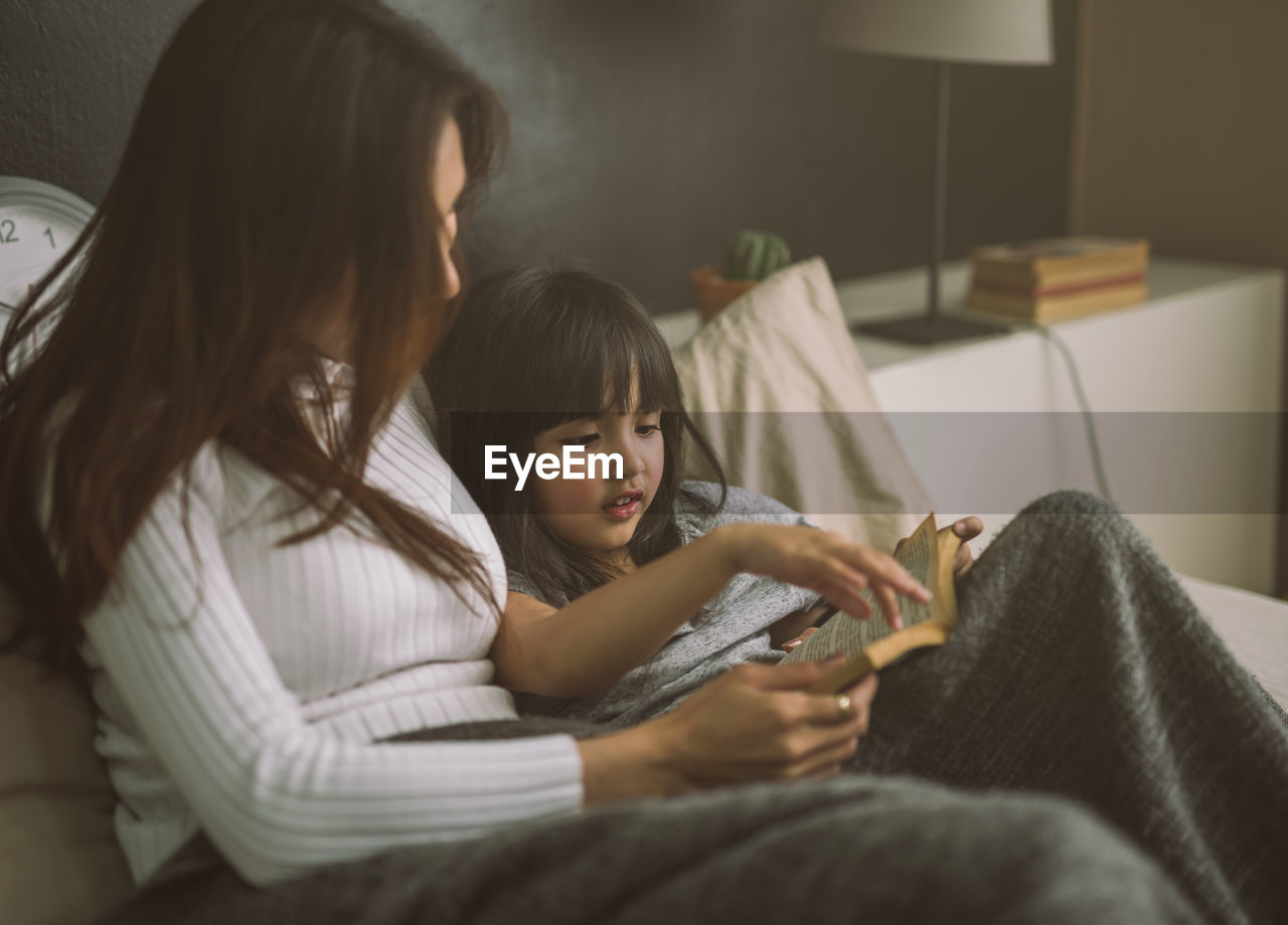 Mother reading book for daughter on bed at home