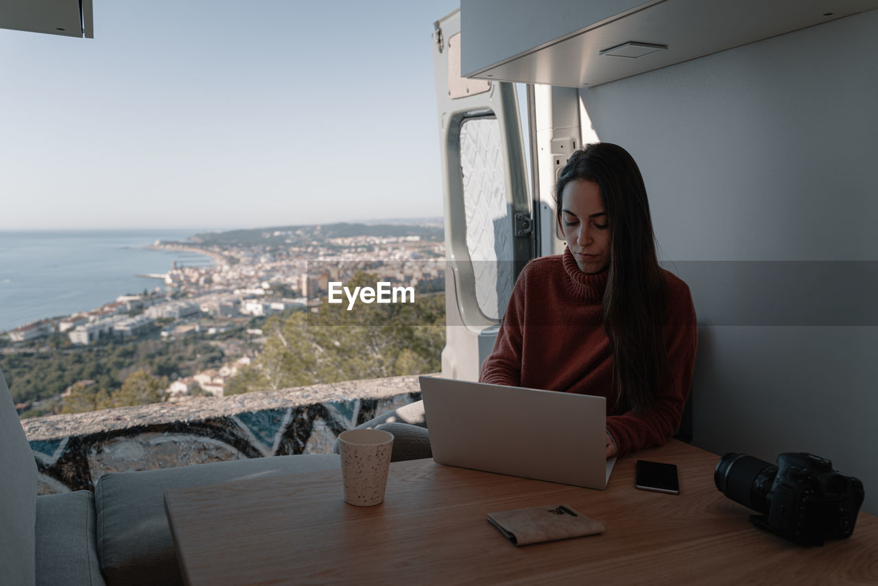 Young woman inside vehicle interior using laptop while sitting on table by sea