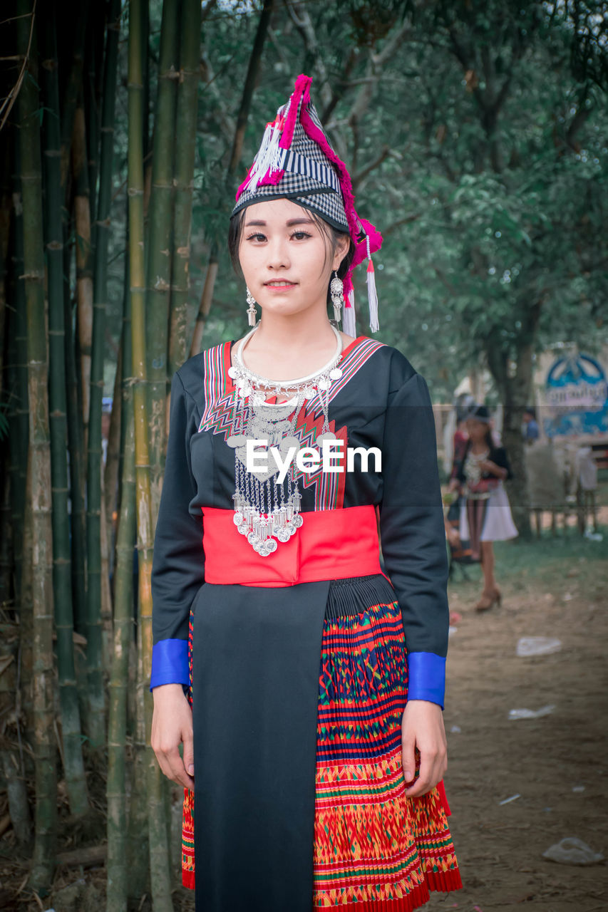 PORTRAIT OF YOUNG WOMAN STANDING AGAINST TREES