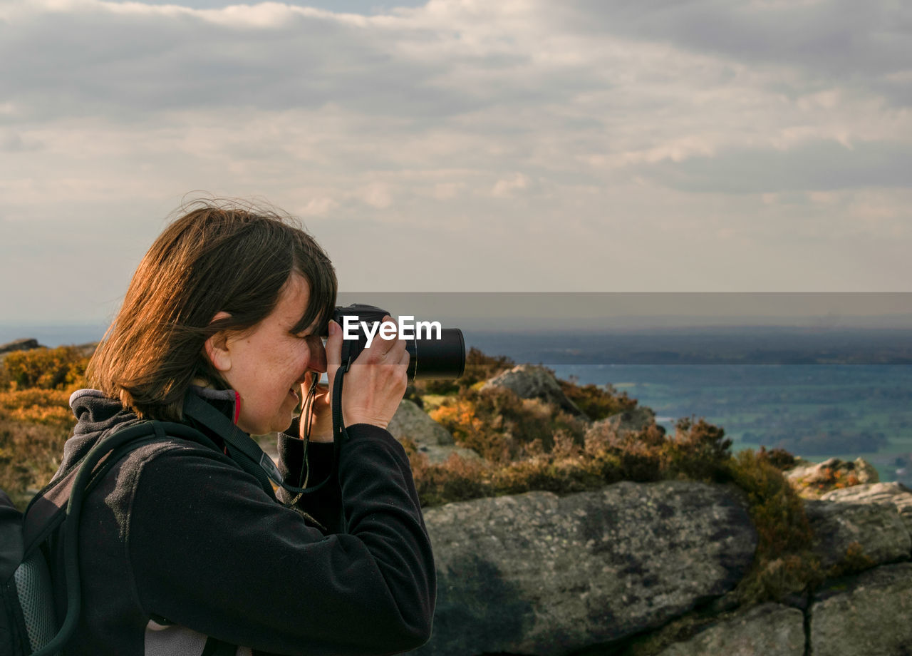 Side view of woman photographing with camera while standing on mountain against cloudy sky during sunset