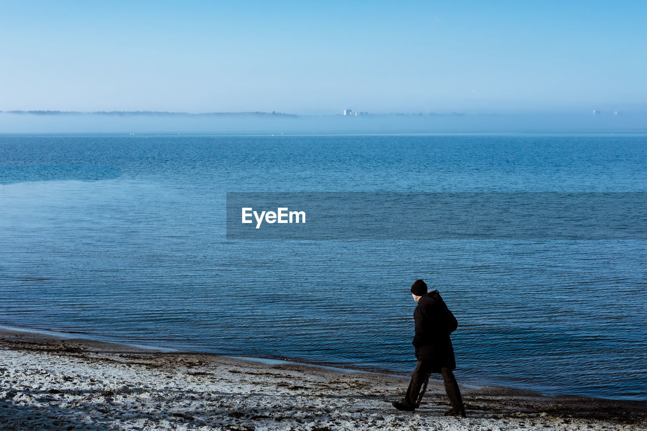 MAN STANDING ON SEA AGAINST CLEAR SKY