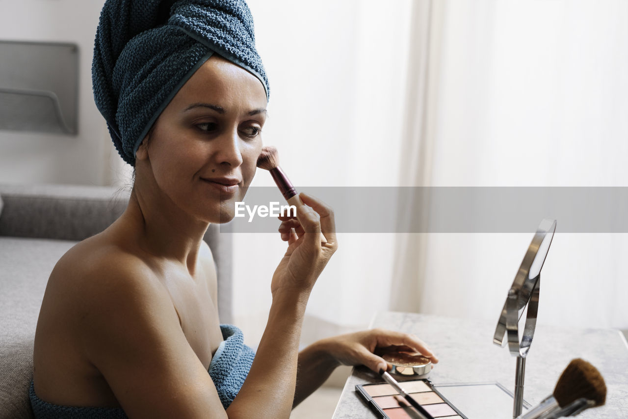 Positive young female wrapped in towels applying powder with brush while doing makeup after shower at home