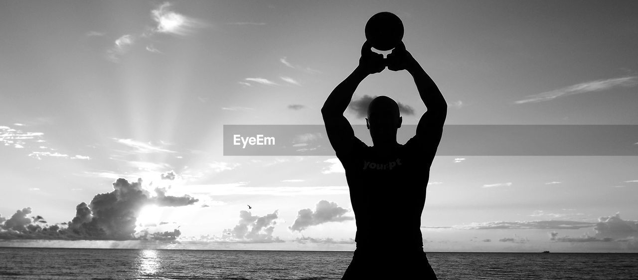 Silhouette man holding kettlebell at beach against sky during sunset