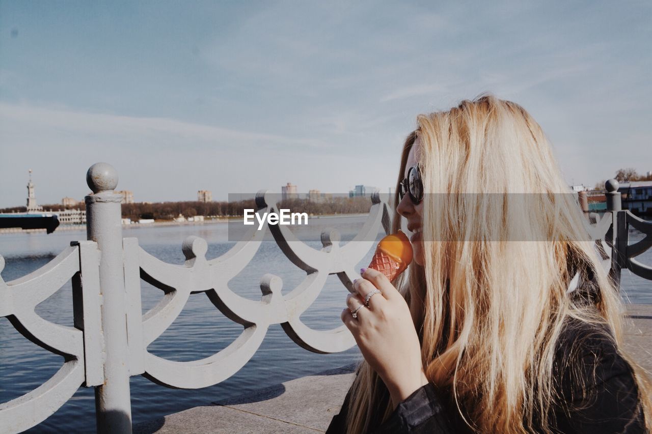 Side view of smiling young woman eating ice cream on pier by sea against sky