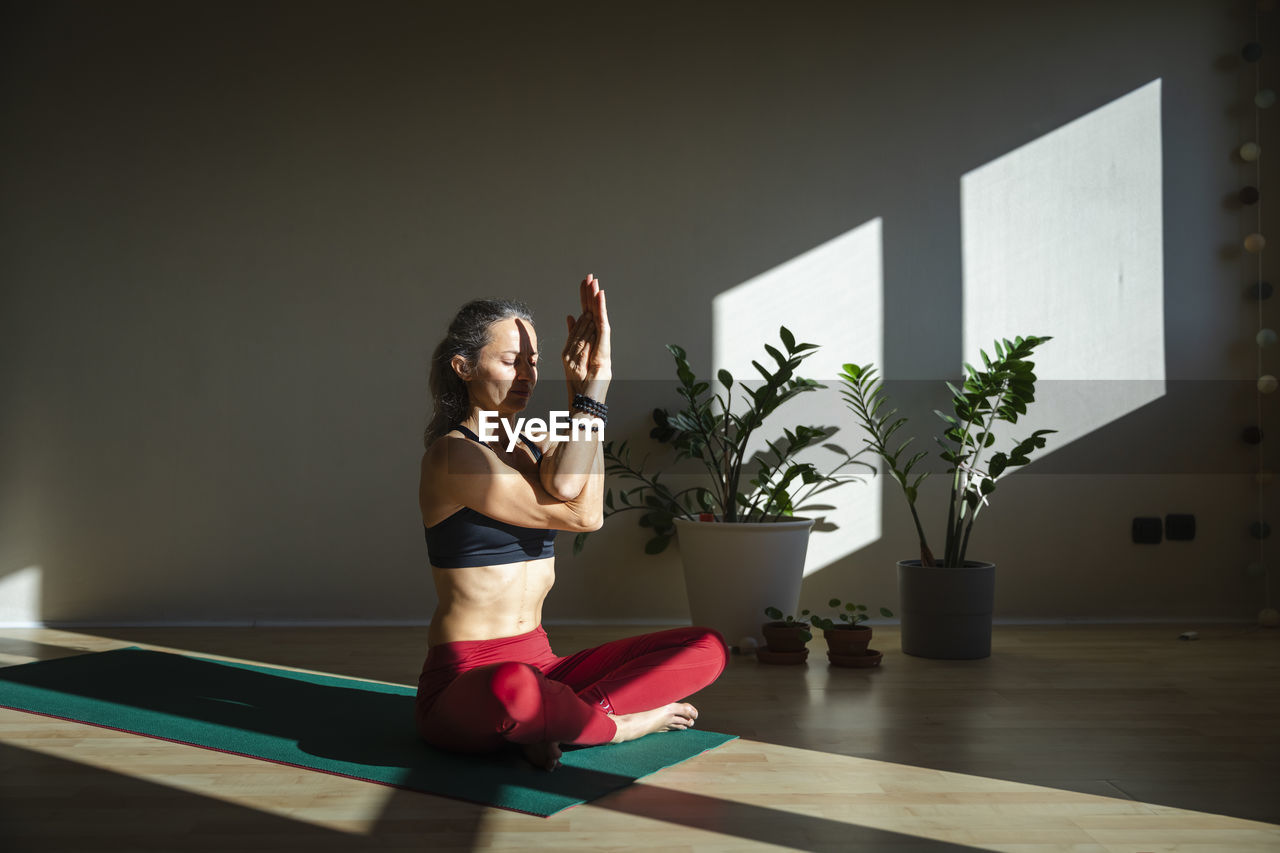 Mature woman with cross legged practicing yoga on mat at home during sunny day