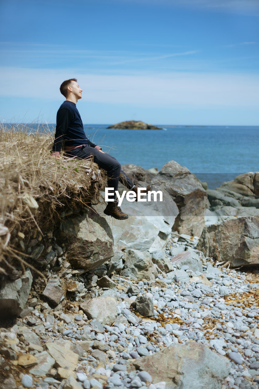 Man sitting on rock by sea against sky