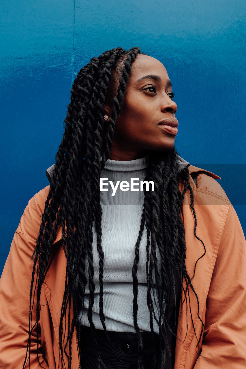 Thoughtful young woman wearing overcoat while standing against blue wall