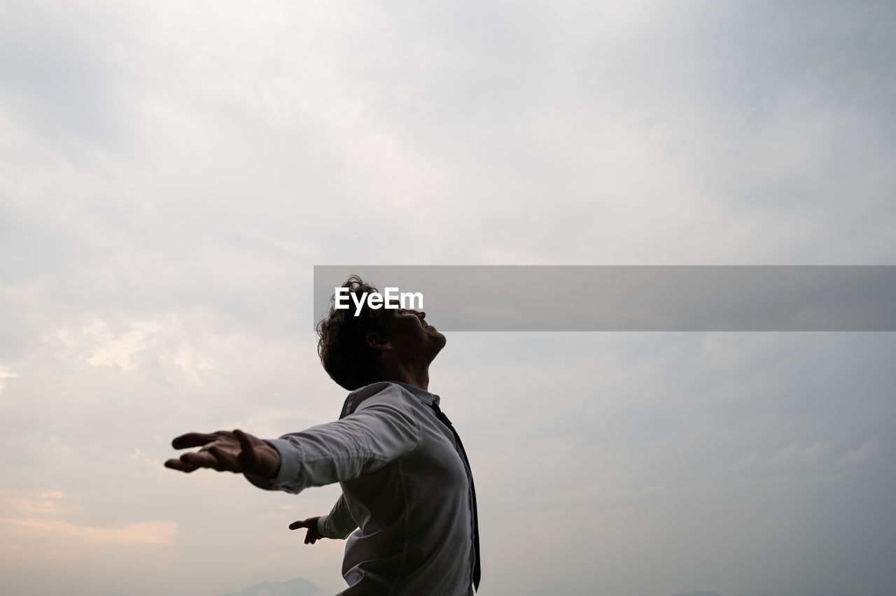 Side view of man with arms outstretched standing against sky