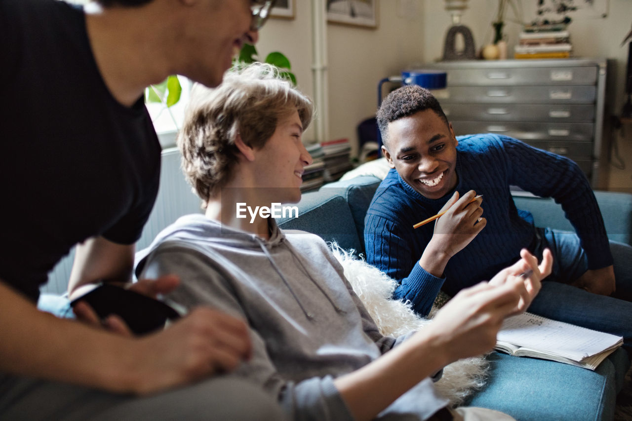 Smiling teenage boy showing mobile phone to friends on sofa while studying at home