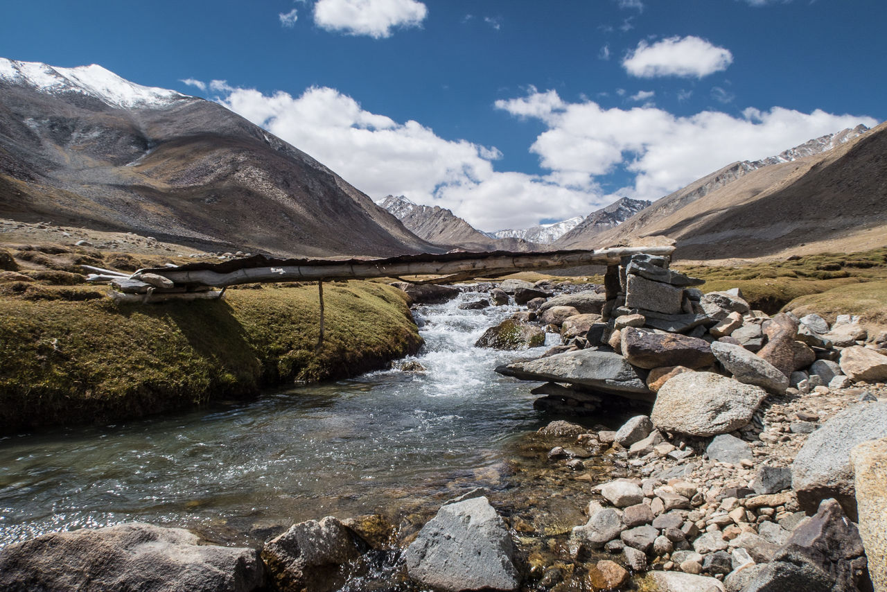 Wooden bridge over river by mountains against cloudy sky