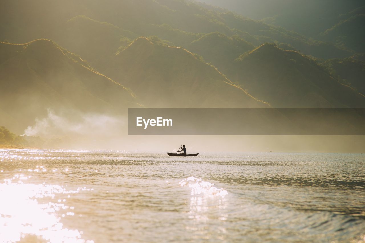 Silhouette man rowing boat on river against mountains