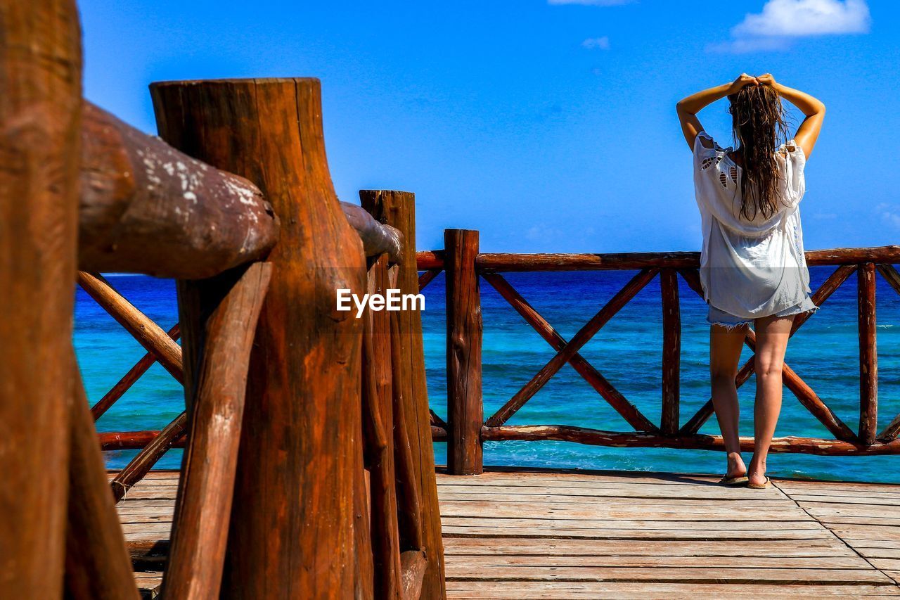 Rear view of young woman with hand in hair standing on pier over sea against blue sky