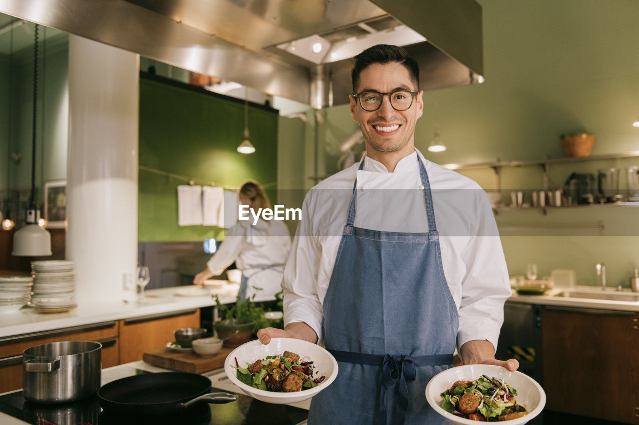 Portrait of confident male chef showing food while standing in commercial kitchen