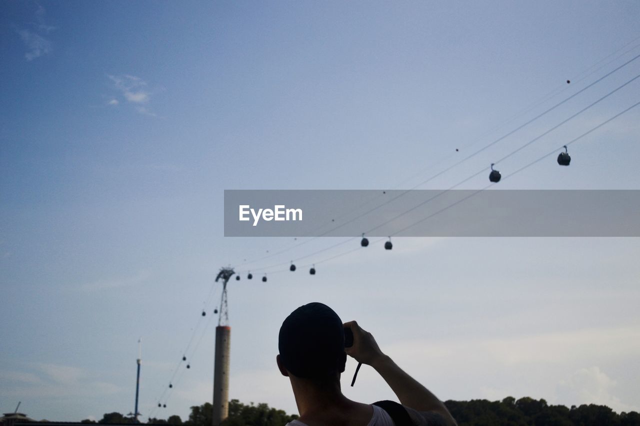 Low angle view of man photographing overhead cable cars against sky