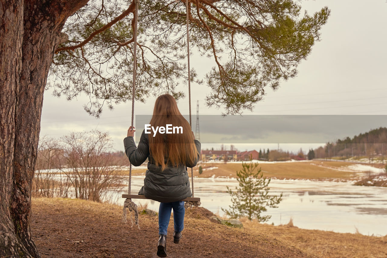 A young woman long blond hair on a wooden swings on a tree branch with icy river on the background..