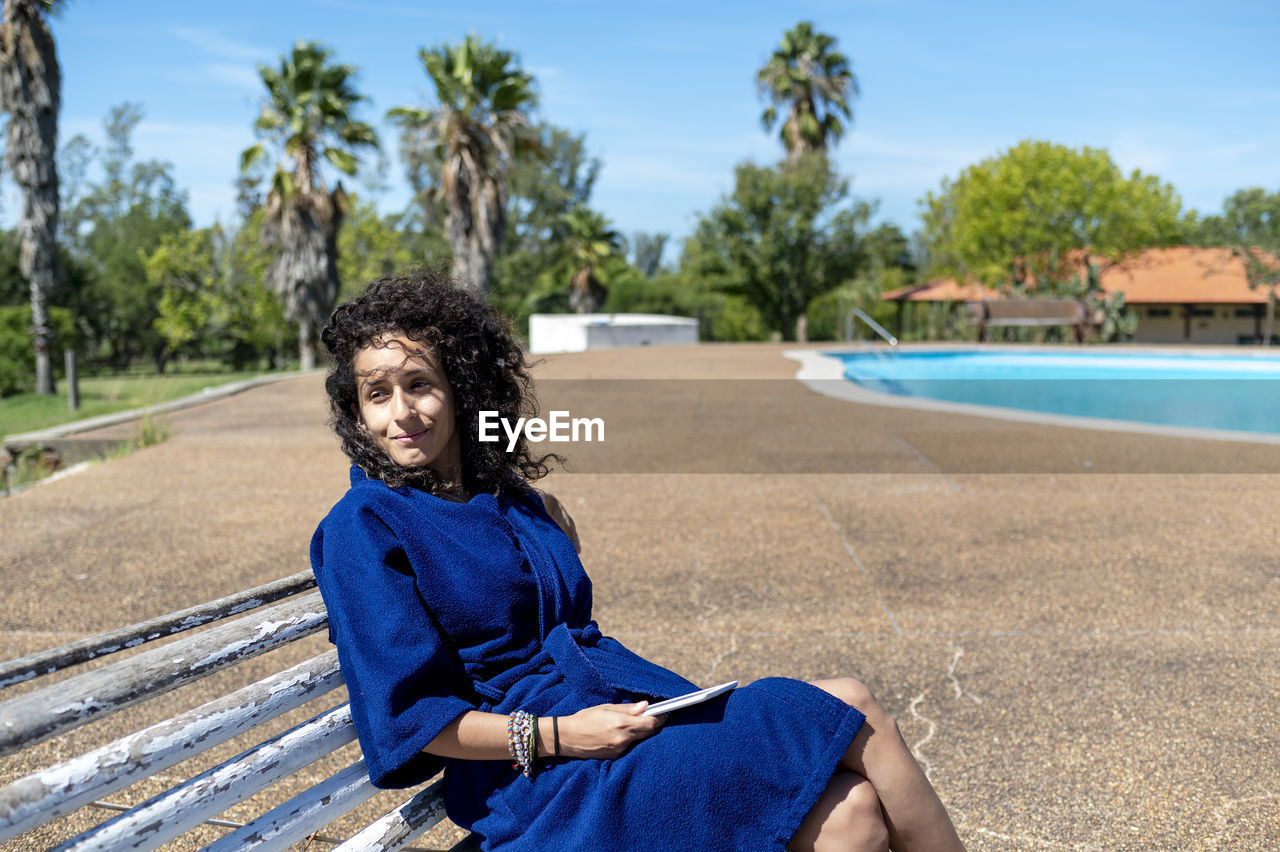 Woman relaxing sitting on a bench outdoors near swimming pool.