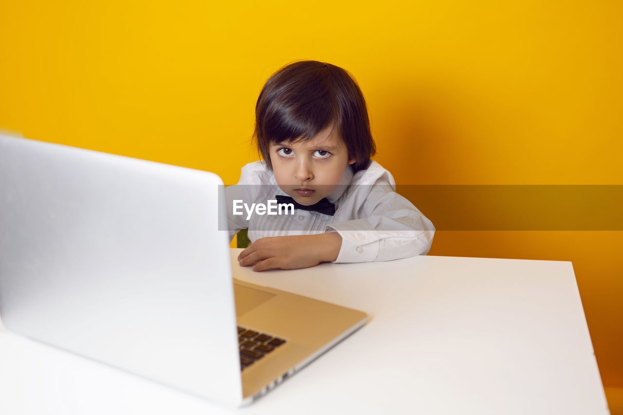 Serious boy child businessman in a white shirt and bow tie sits at a laptop desk in a yellow office