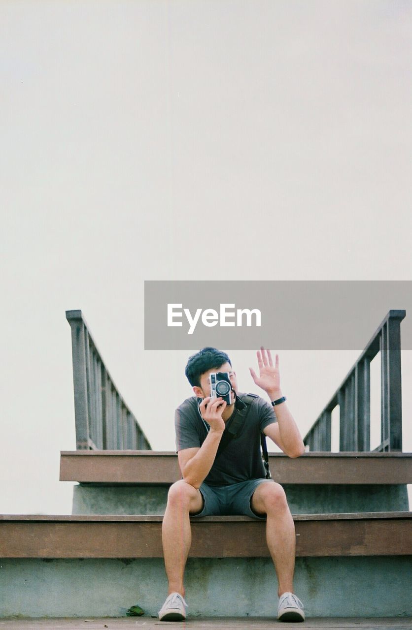 Man photographing with camera on steps while waving against clear sky