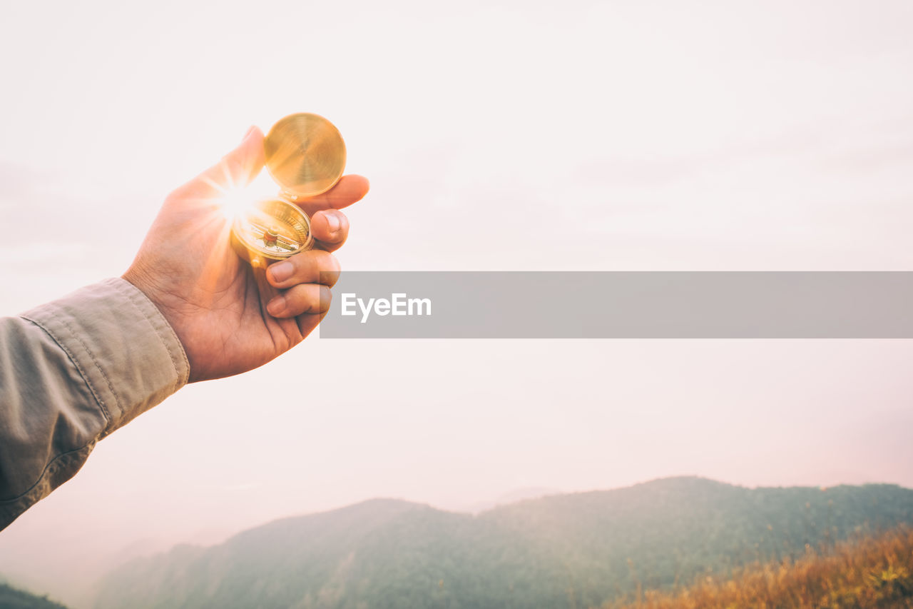 Close-up of hand holding compass against sky during sunset