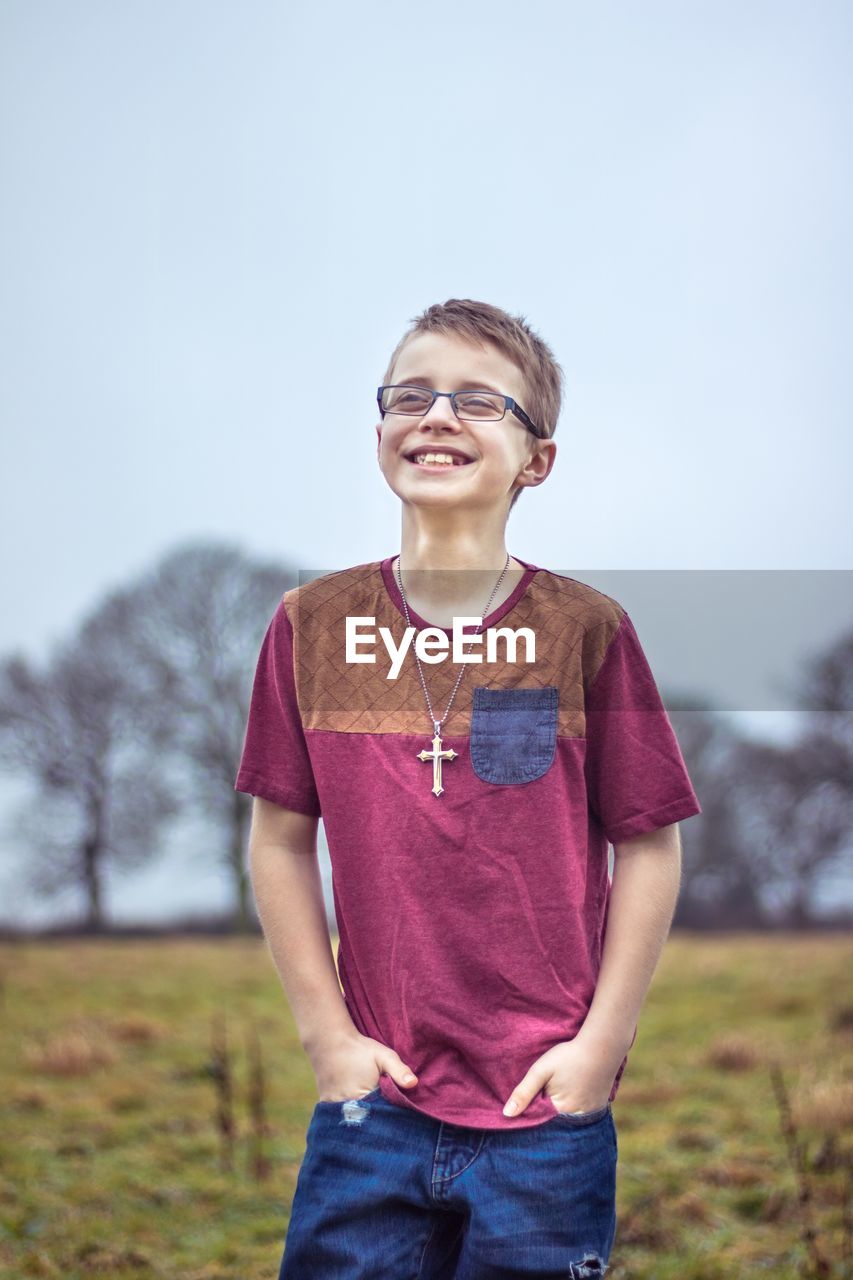 Smiling boy standing with hands in pockets on field against clear sky