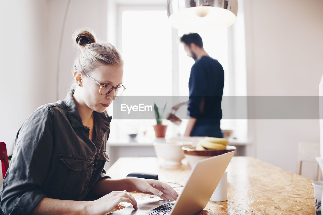 Woman working on laptop against man watering plant at home