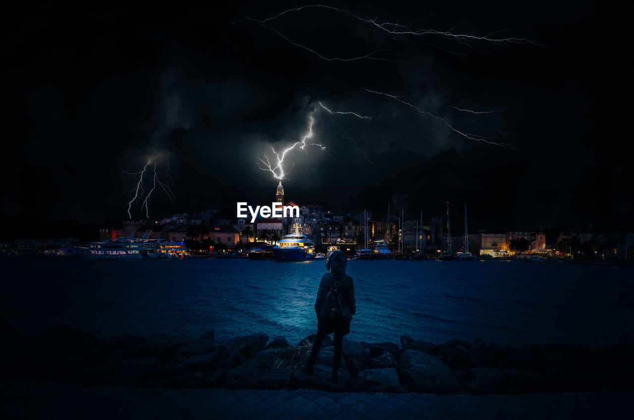 Panoramic view of illuminated city by sea against thunder sky at night