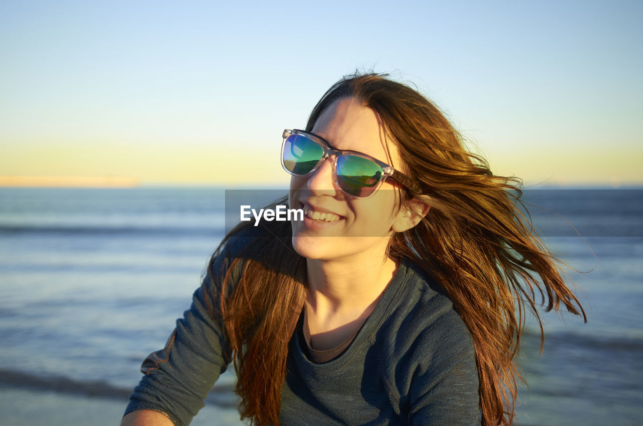 Smiling young woman wearing sunglasses at beach against sky during sunset